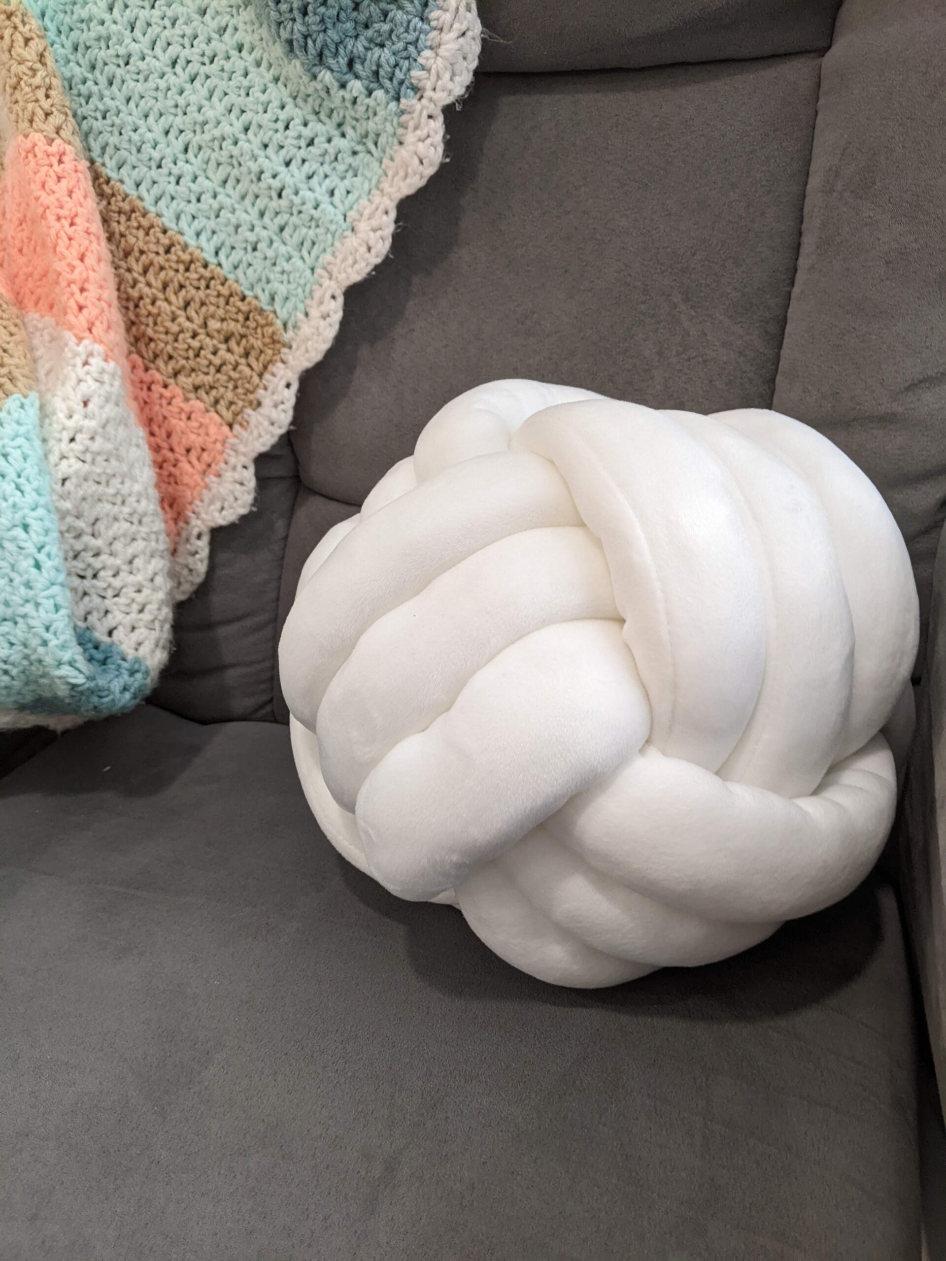 Soft white knot pillow ball on a gray couch with a crocheted blanket