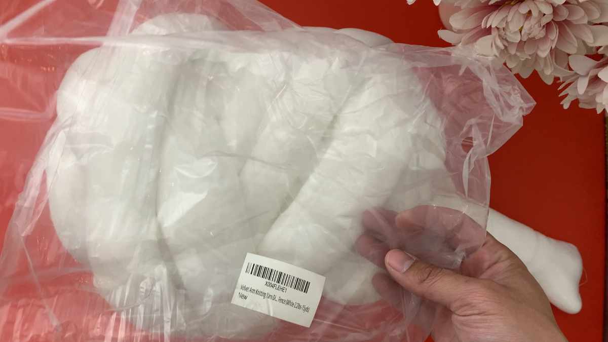 Jumbo tube yarn in its original plastic packaging with a barcode label, held by a hand against a red background with pink flowers.